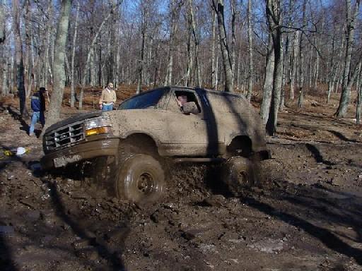 Mud-covered two-door Ford Bronco stuck in deep mud in bare woods, driver visible inside.