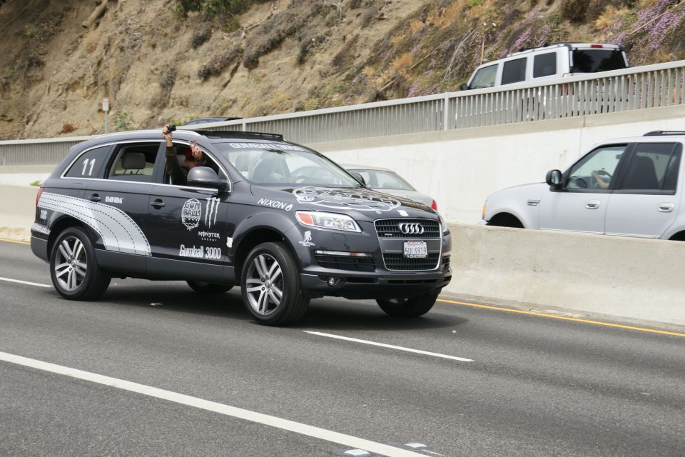 Dark Audi Q7 with Monster Energy and Gumball 3000 decals driving on highway. Person in driver's seat holding phone