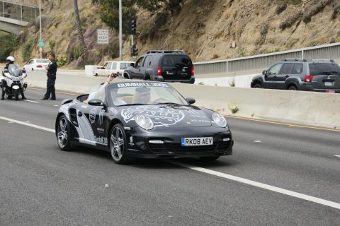1993 Ford Taurus - A black Porsche 911 convertible with Gumball 3000 livery drives on the highway, with police presence.