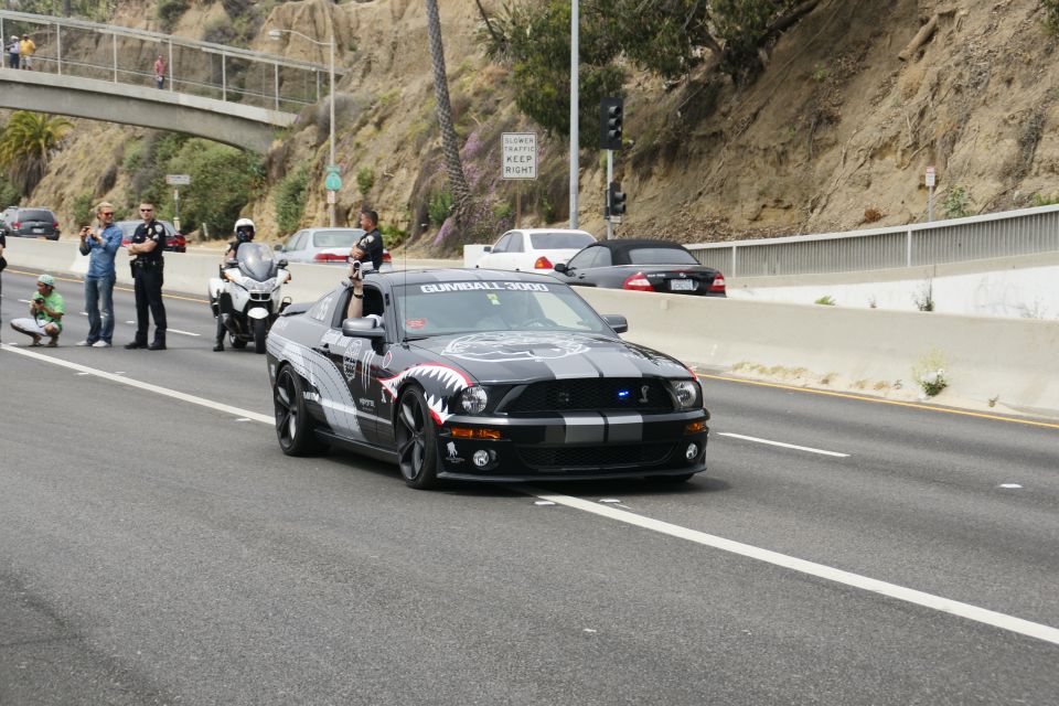 Black Ford Mustang with shark teeth graphics and Gumball 3000 text on windshield, driving on highway