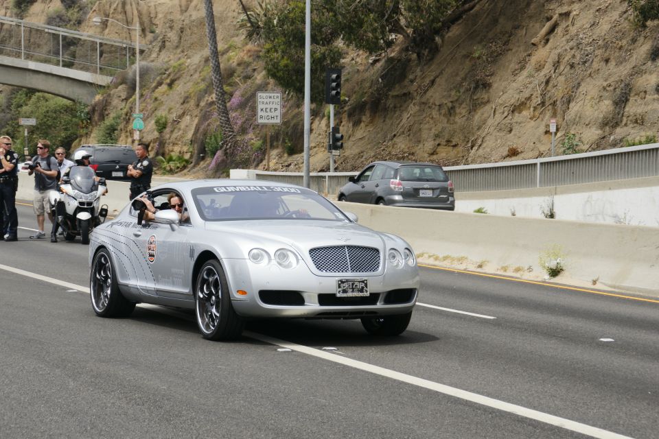 Silver Bentley Continental GT with Gumball 3000 sticker on windshield, driving on highway with police motorcycle nearby