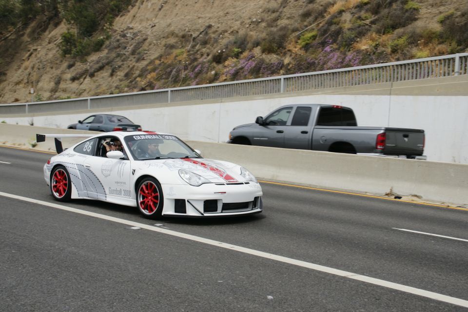 White Porsche 911 with red accents and racing decals, number 86, on highway. Gray pickup truck and dark sedan also visible