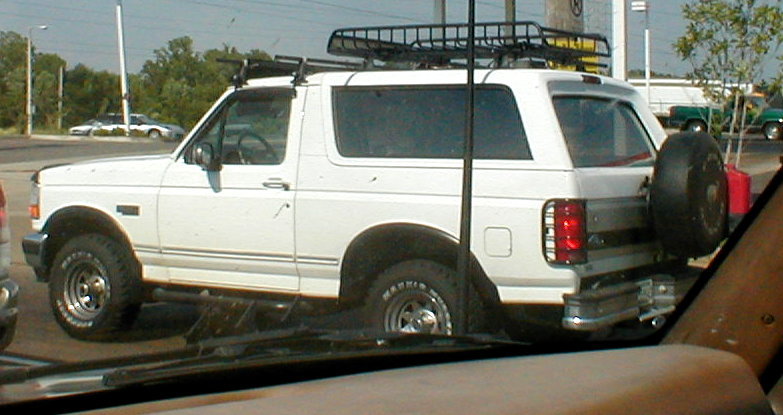 White two-door Ford Bronco SUV with roof rack basket and rear spare tire, side-rear view