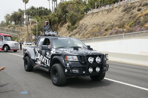 1993 Ford Taurus - A heavily customized black pickup truck with a person standing in the bed drives down a road, accompanied by another vehicle.