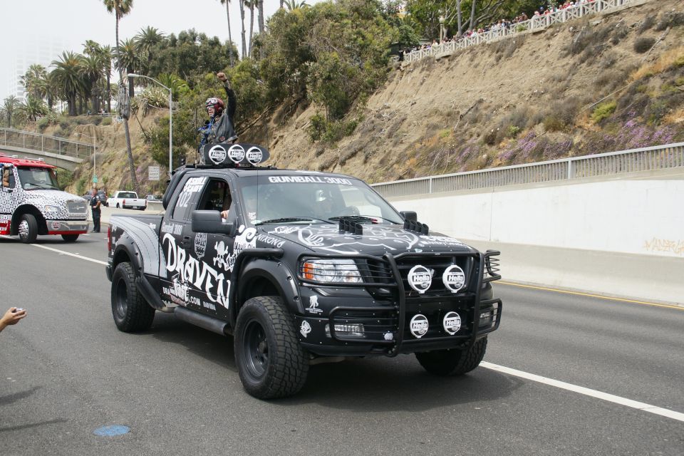 Black pickup truck with 'DRAVEN' decals and Gumball 3000 text on windshield, driving on a road with a person standing in the bed