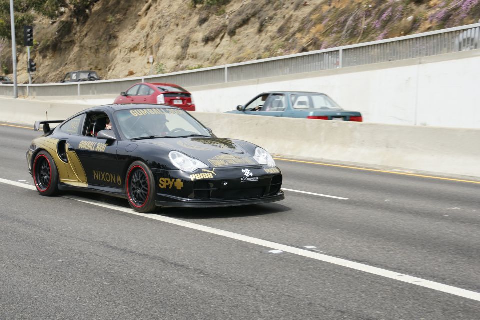 Black Porsche 911 with gold accents and Gumball 3000 livery, driving on a highway