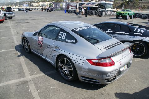 1993 Ford Taurus - A silver Porsche 911 and a black Lamborghini are parked in a lot, both displaying racing numbers and sponsor decals.