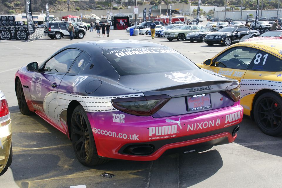 Rear view of a Maserati GranTurismo with a custom pink, purple, and black livery, parked next to a yellow race car
