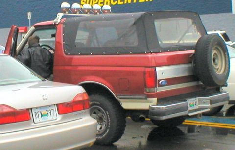 1983 Ford Bronco - A red Ford Bronco with a soft top and a spare tire mounted on the rear is parked next to another vehicle.