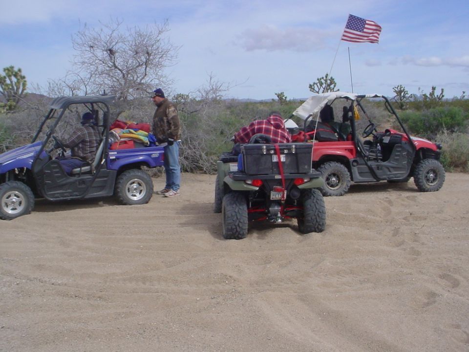 Three ATVs parked on dirt in a desert landscape. One blue, one red, and one green. An American flag flies from the red ATV