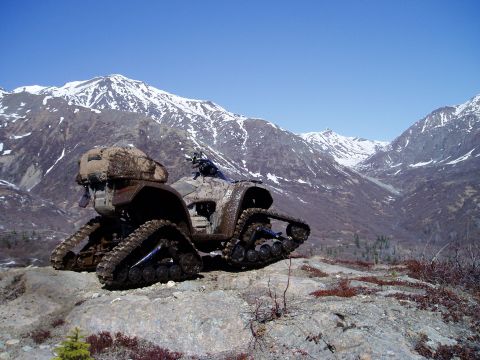 2000 Ford E350 - A muddy ATV with tracks instead of wheels is parked on a rocky slope with snow-capped mountains in the background.