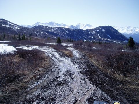 2000 Ford E350 - A muddy, rutted dirt road winds through a landscape with sparse vegetation and snow-capped mountains in the background under a clear blue s…
