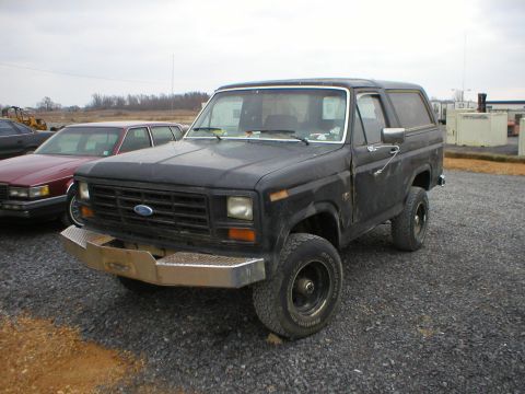1983 Ford Bronco - This black Ford Bronco is parked outdoors with other vehicles and structures visible.