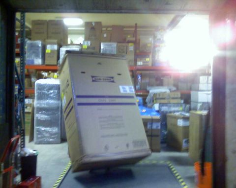 2006 Ford E350 - A large cardboard box is tilted on a pallet jack in a warehouse setting. The box has purple text and labels on it, and there are shelves of…