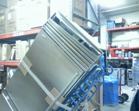 2006 Ford E350 - A large stainless steel appliance, possibly a refrigerator or freezer, is tilted on a blue hand truck in a warehouse setting. Cardboard box…