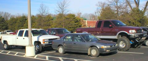 1983 Ford Bronco - A collection of trucks and a sedan are parked in a lot, with one lifted truck oversized tires.