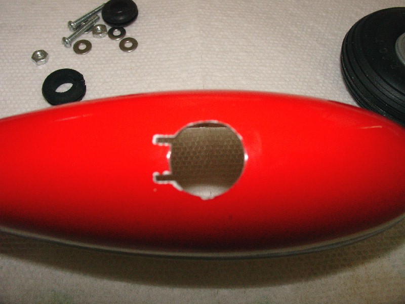 Red fuselage part with a circular opening and two small tabs, surrounded by screws, nuts, and washers