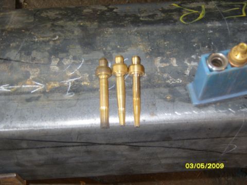 1989 Ford Bronco - Three brass welding torch tips are shown resting on a metal surface, with a blue plastic holder containing additional parts visible on the…