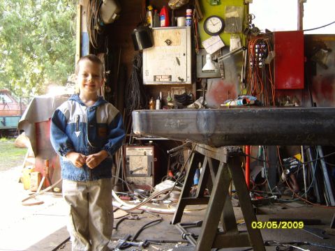 1989 Ford Bronco - A young boy stands in a cluttered workshop, with a large metal fabrication project in the foreground.