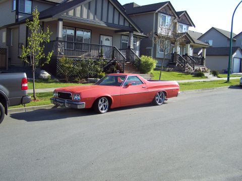 1973 Ford Ranchero - Red Ford Ranchero coupe utility parked at the curb of a suburban street lined with two-story houses, seen from the driver's side front thre…