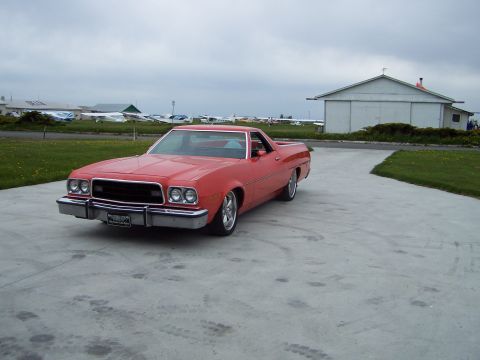 1973 Ford Ranchero - Red Ford Ranchero coupe utility on a concrete apron at a small airfield, with parked light aircraft and a metal hangar behind it under an o…