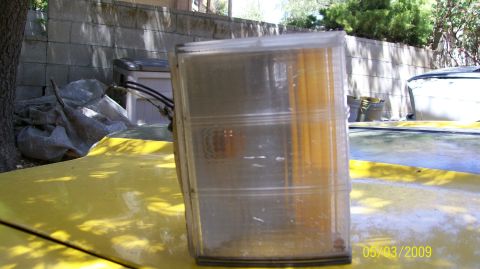 1972 Ford Ranchero - A close-up of a headlight assembly resting on a yellow surface, with a wall and trees in the background.