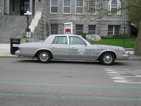 1991 Ford LTD Crown Victoria - Side view of a silver Rhode Island State Police sedan with roof beacon parked at a curb before a stone government building.