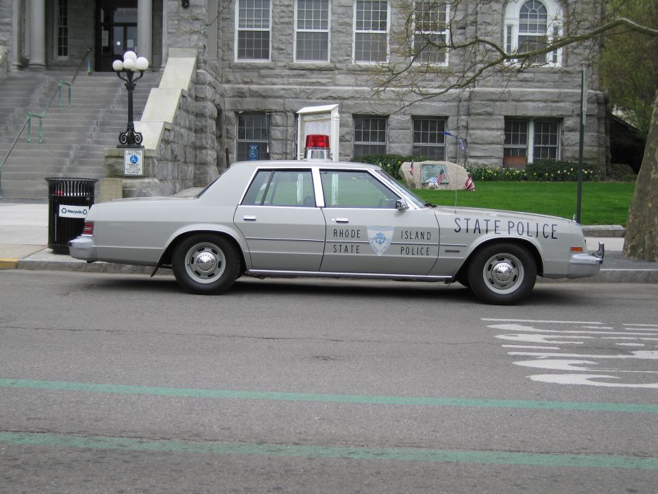 Silver Rhode Island State Police sedan parked at a curb by a stone building
