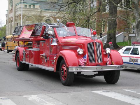 1991 Ford LTD Crown Victoria - vintage red fire engine with a long ladder and American flags drives in a parade past a stone building and a school bus.