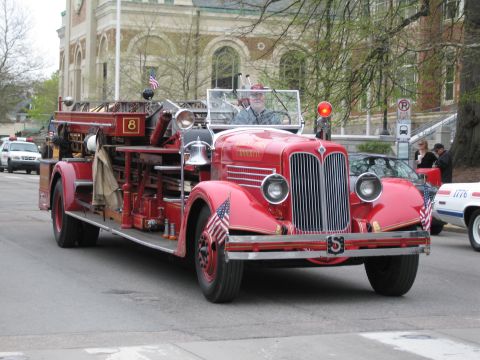 1991 Ford LTD Crown Victoria - antique red open-cab fire engine marked with the number 8 and American flags drives in a parade past a stone building.