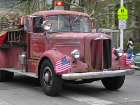 1991 Ford LTD Crown Victoria - weathered red antique fire engine with American flags and a roof beacon drives along a street during a parade.