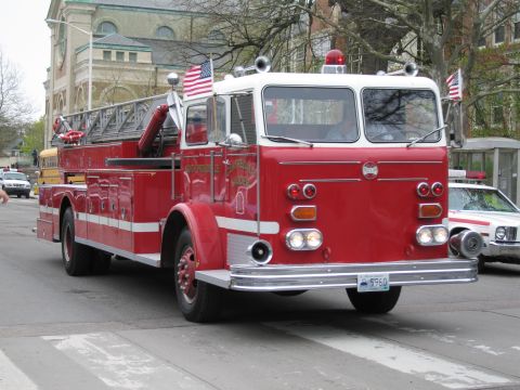 1991 Ford LTD Crown Victoria - large red-and-white aerial-ladder fire truck with an American flag drives in a parade past buildings and a police car.