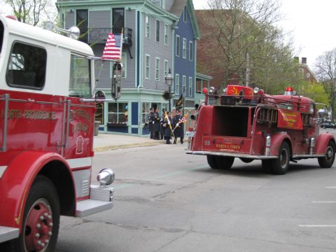 1991 Ford LTD Crown Victoria - Two vintage red fire engines pass on a town street with an American flag and ornate Victorian buildings in the background.