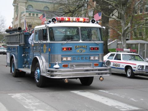 1991 Ford LTD Crown Victoria - blue Hahn fire engine lettered Engine 7 rolls down a street with flags on its roof and a white-and-red police wagon following.