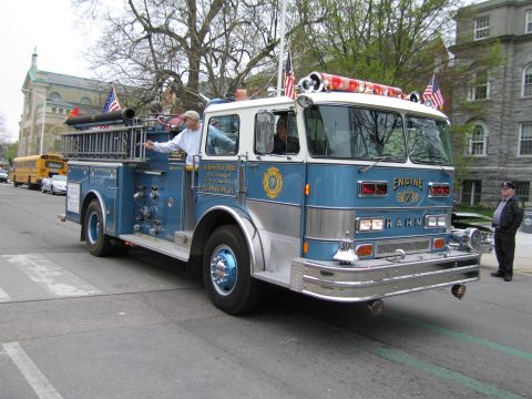 1991 Ford LTD Crown Victoria - blue Hahn fire engine marked Engine 7 drives past stone buildings as a man rides the running board and a yellow school bus waits behind.