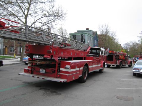 1991 Ford LTD Crown Victoria - Rear three-quarter view of a red aerial ladder fire truck with its long ladder stowed on top, followed by other emergency vehicles.