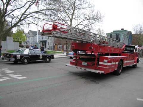 1991 Ford LTD Crown Victoria - red aerial ladder fire truck and a black-and-white police sedan travel a street lined with bare trees and houses.