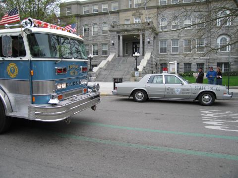 1991 Ford LTD Crown Victoria - blue Hahn fire engine sits beside a silver Rhode Island State Police sedan in front of a large stone institutional building.