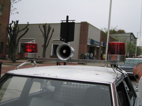 1991 Ford LTD Crown Victoria - Detail of a white car roof fitted with twin red rotating beacon lights and a chrome public-address speaker, with storefronts behind.