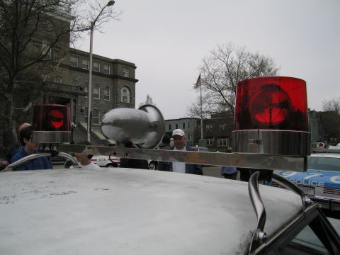 1991 Ford LTD Crown Victoria - Roof of a white car fitted with two red rotating beacons and a chrome siren, with onlookers and stone buildings in the background.