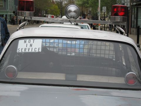 1991 Ford LTD Crown Victoria - Back window of a vintage police car displaying a Not In Service sign and a prisoner cage, topped with red beacons and a spotlight.