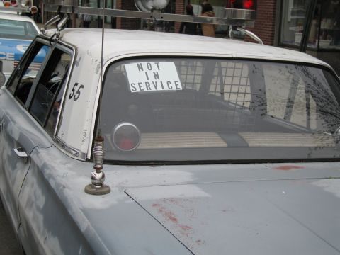 1991 Ford LTD Crown Victoria - Rear quarter of a weathered silver police car marked 55 with a Not In Service sign in the rear window and a whip antenna on the fender.