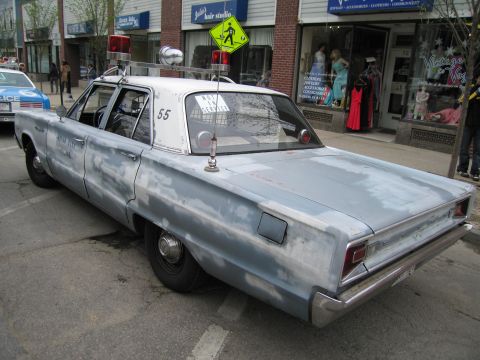 1991 Ford LTD Crown Victoria - primered silver four-door classic police sedan marked 55 with roof beacons parks along a street lined with shops.