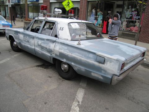 1991 Ford LTD Crown Victoria - Rear three-quarter view of a primered silver four-door classic police sedan marked 55 parked diagonally on a shopping street.