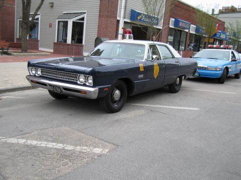 1991 Ford LTD Crown Victoria - dark blue classic Plymouth state-police sedan with a single roof beacon parks at a corner beside a light-blue modern police car.