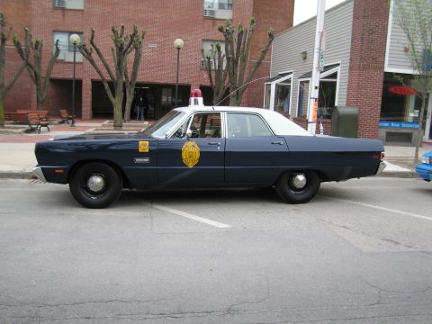 1991 Ford LTD Crown Victoria - Side profile of a dark blue four-door classic state-police sedan with a gold door shield and roof beacon parked at a curb.
