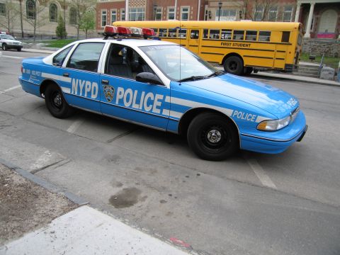 1991 Ford LTD Crown Victoria - light-blue NYPD police car with roof lightbar parks on a sloped street near a First Student yellow school bus.