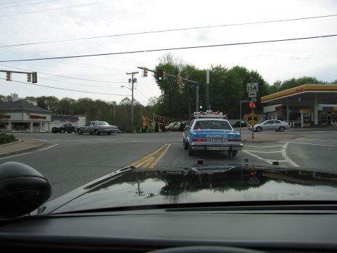 1991 Ford LTD Crown Victoria - Dashboard view through a windshield toward a white police car crossing an intersection, with a green traffic light and gas station.