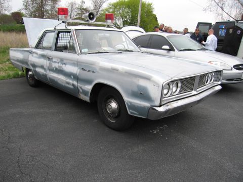 1991 Ford LTD Crown Victoria - weathered light-blue mid-1960s Dodge police sedan with roof beacons and a siren, parked in a lot near a crowd.