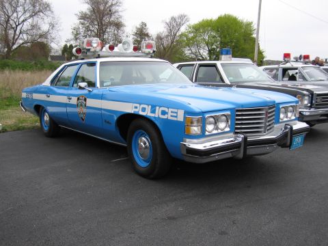 1991 Ford LTD Crown Victoria - blue-and-white police sedan lettered 'POLICE' with roof beacons and blue wheels, parked among other patrol cars.